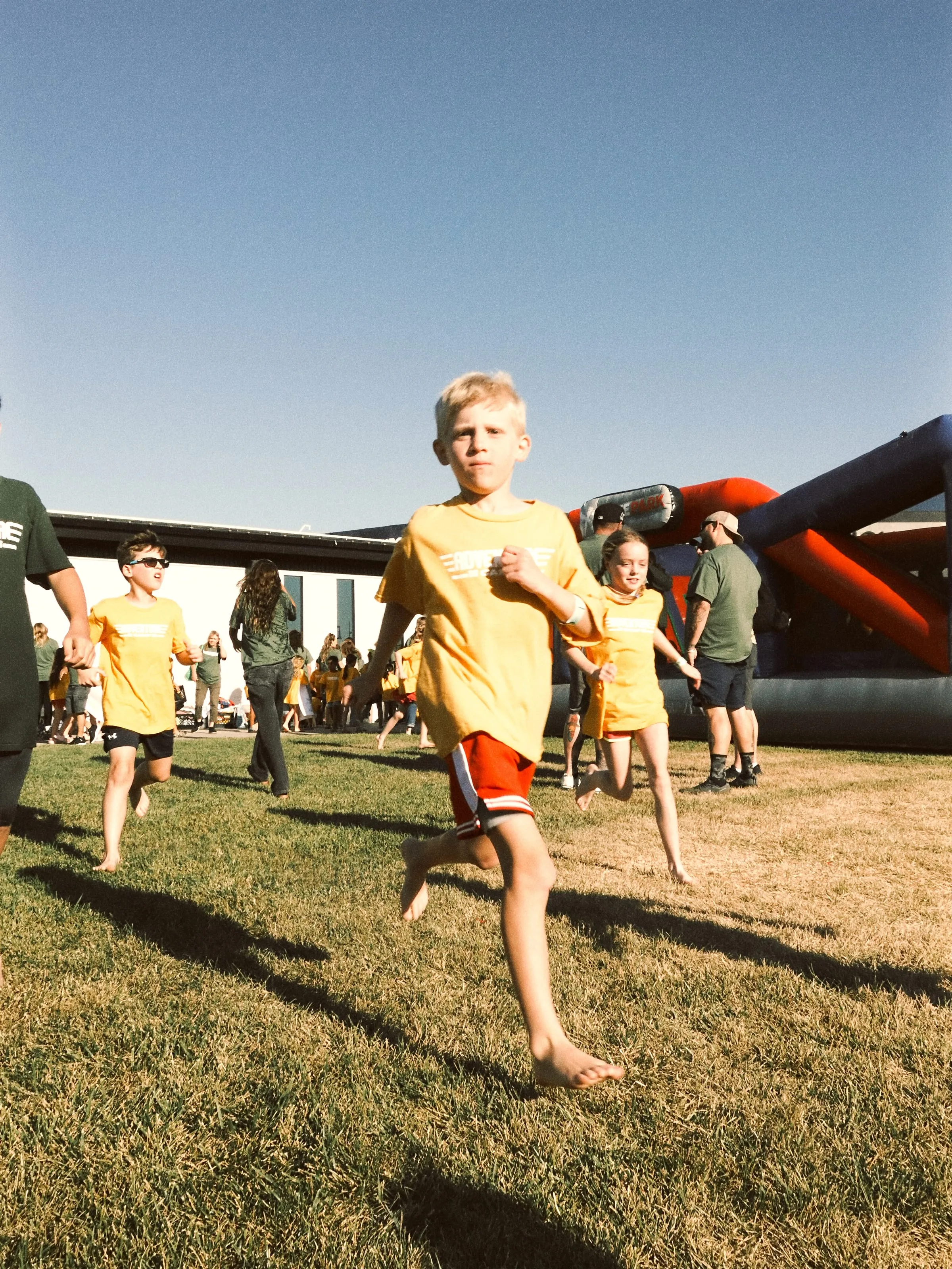 Campers in yellow shirts race barefoot across the lawn past an inflatable obstacle course.