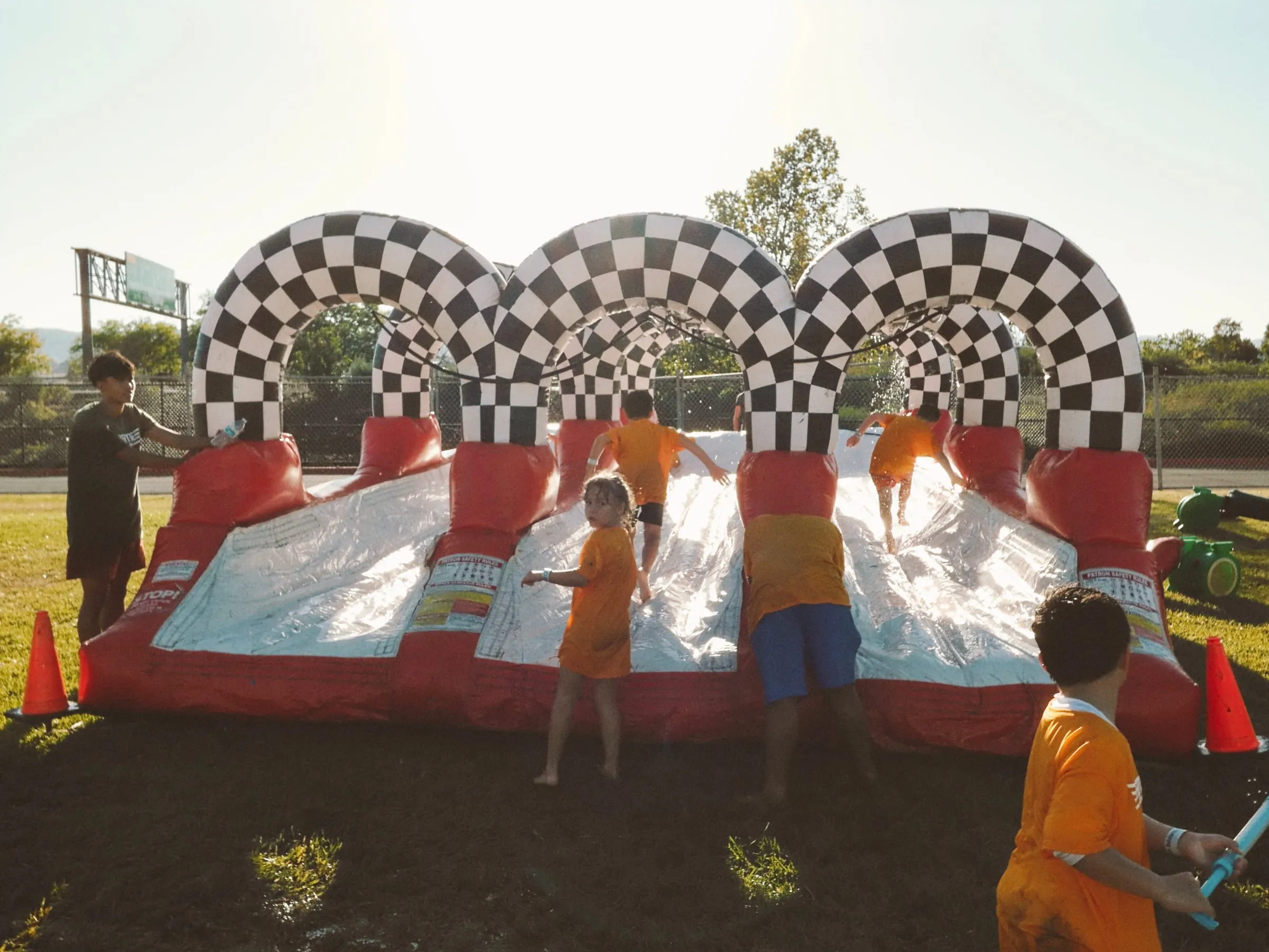 Campers dash through a checkered inflatable slide tunnel in the afternoon sun.