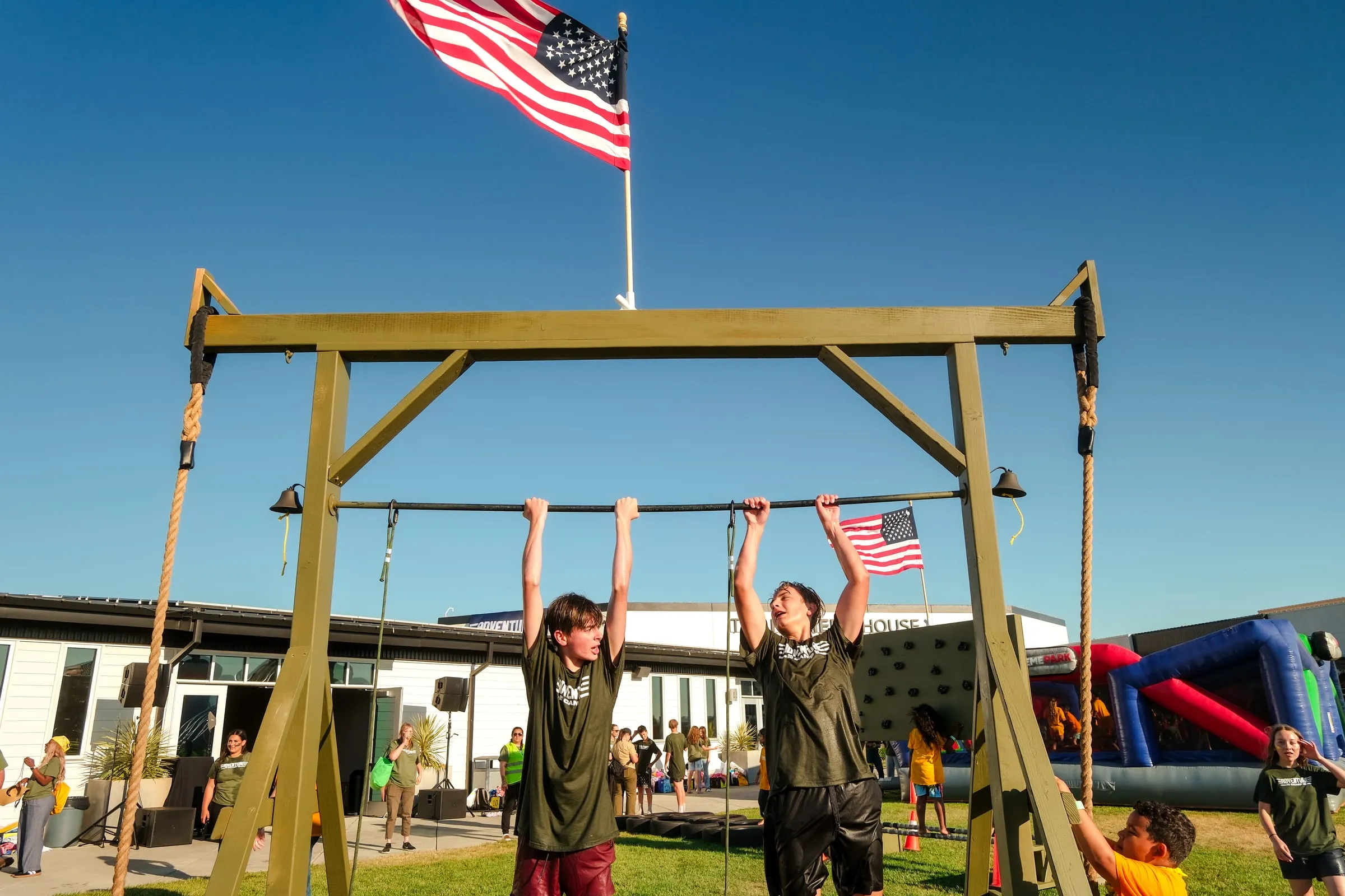 Two campers reach the top bar of the obstacle course as the flag flies overhead.