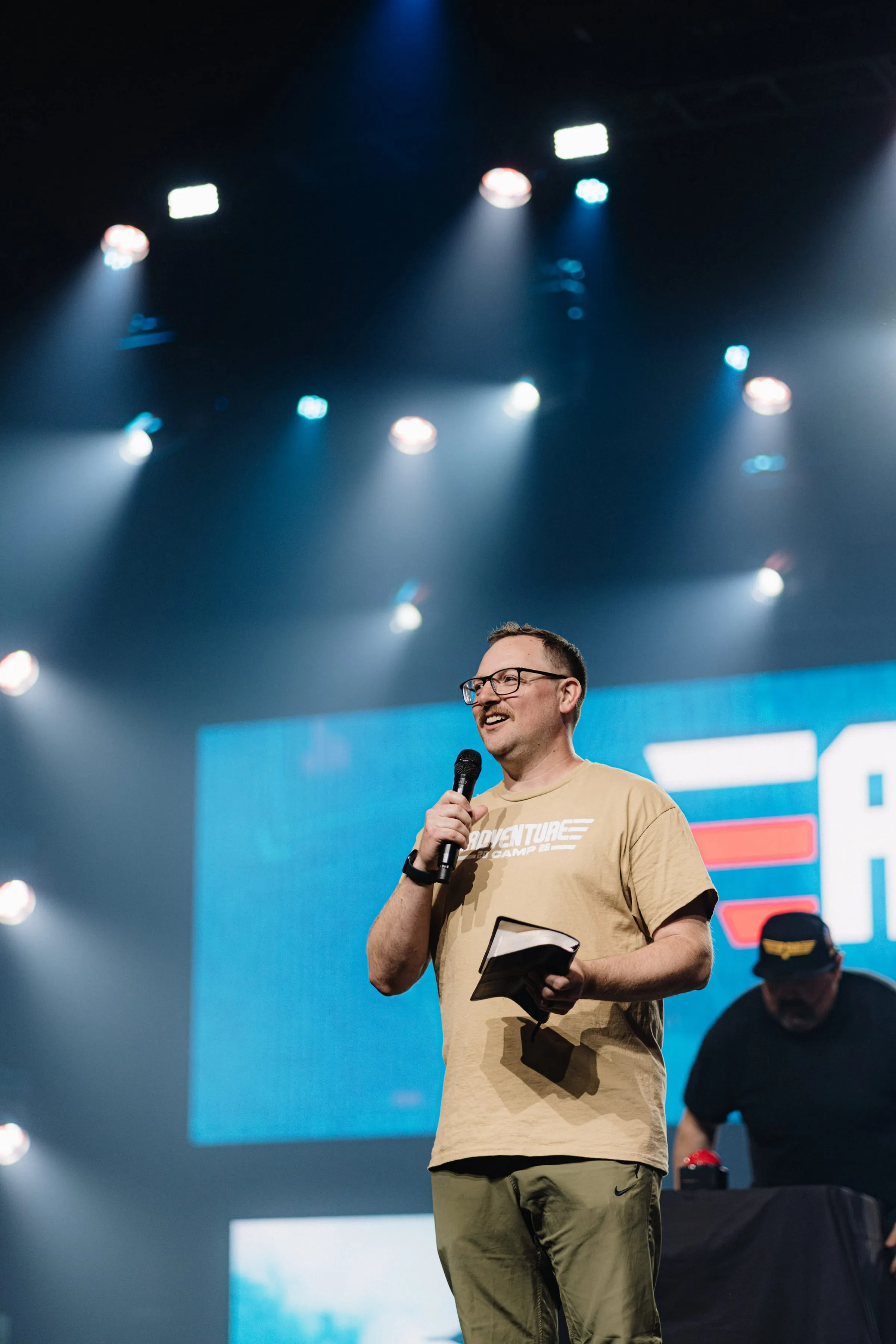A camp host addresses the crowd from the stage, notebook and mic in hand.