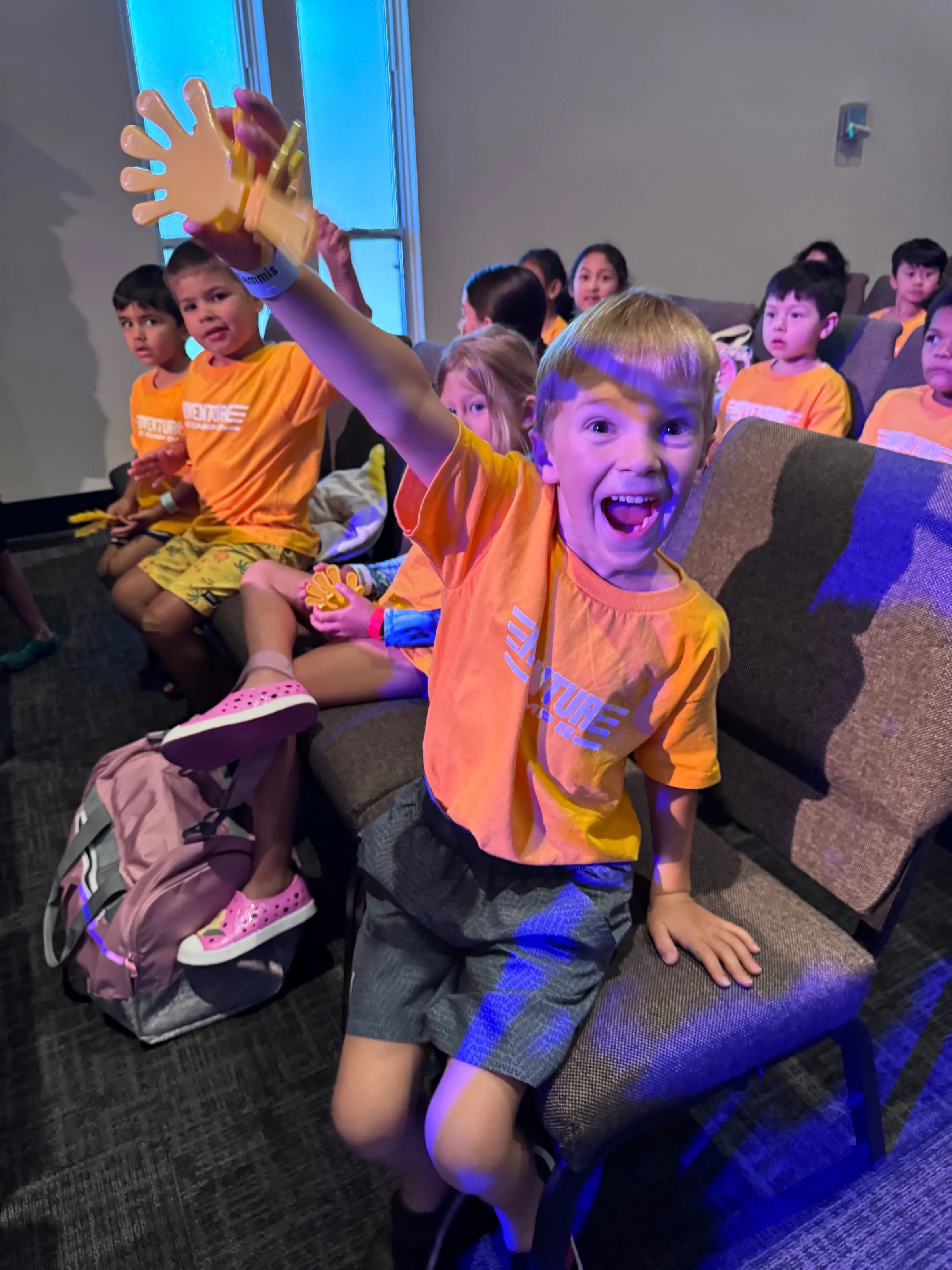 A camper waves a giant foam hand at the camera during a glow-lit evening party.
