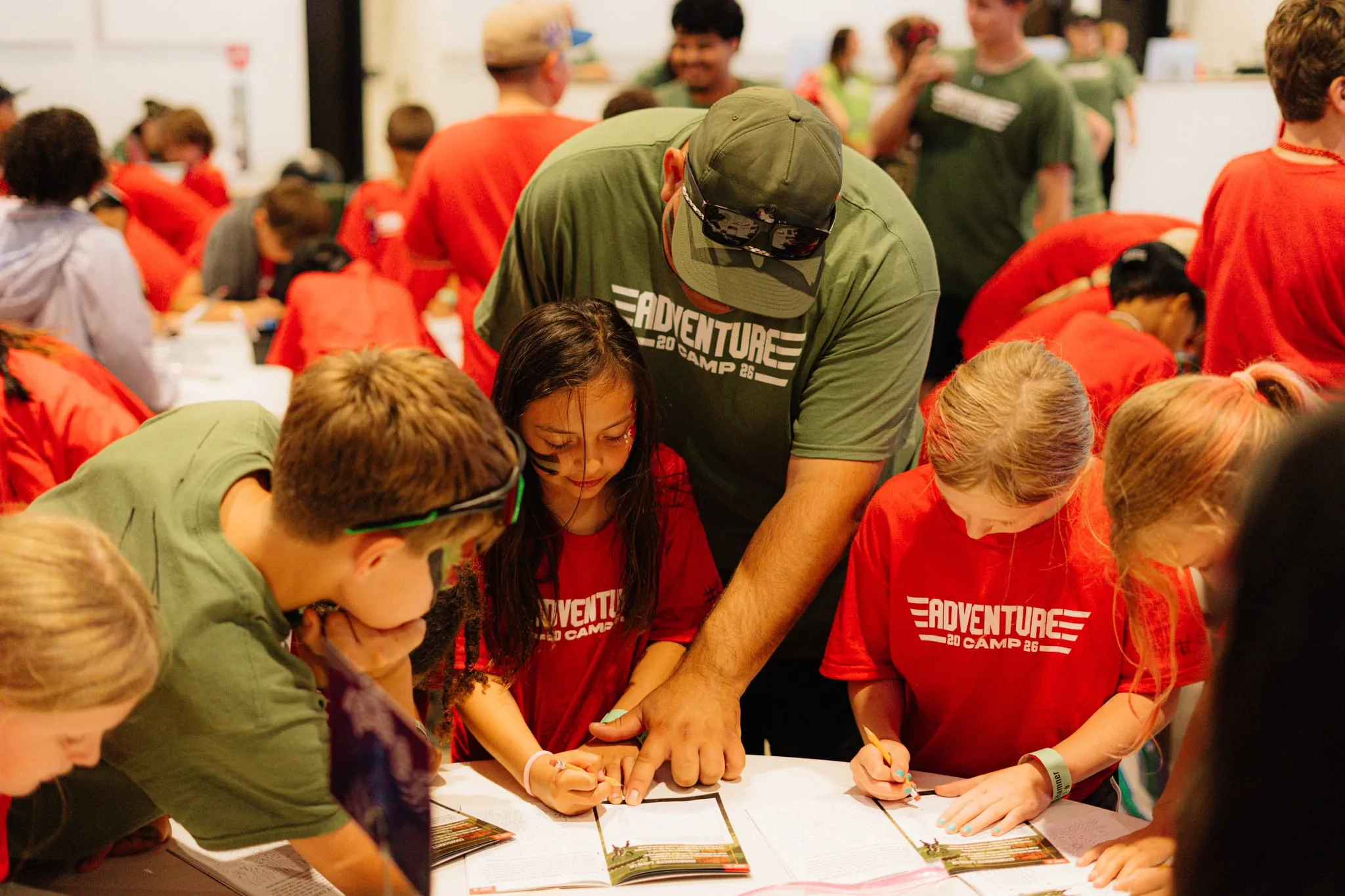 A camp leader helps campers fill out craft sheets at a table.