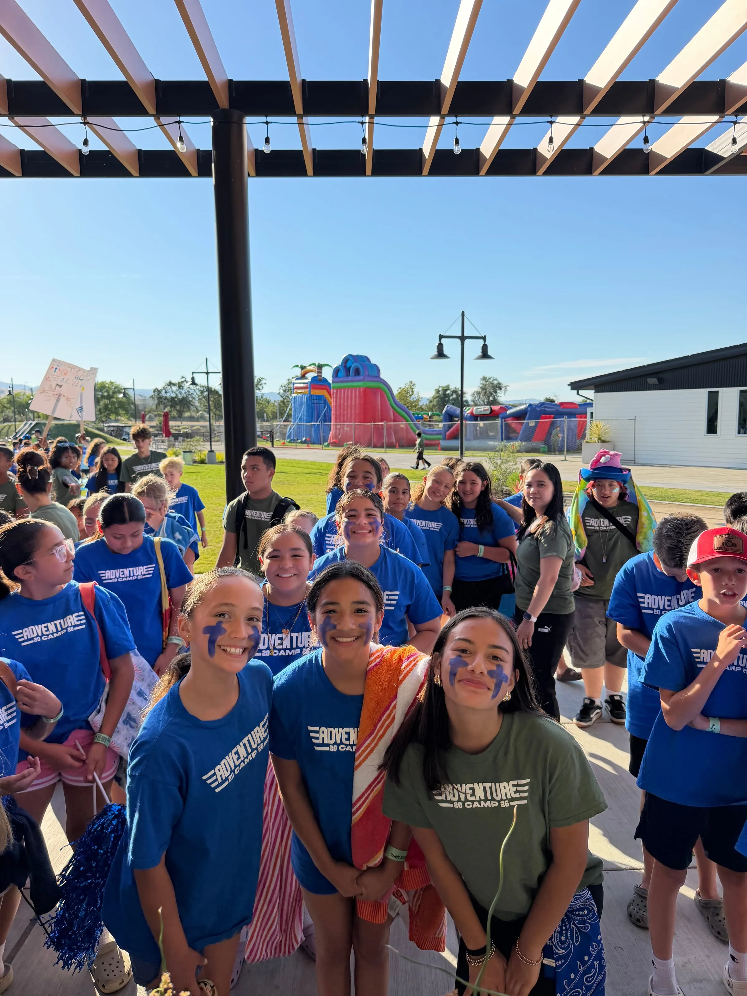 A large group of campers gathers near the string-light pergola after a day of games.